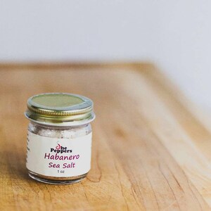 May include: A small glass jar with a gold lid filled with white sea salt. The label reads "The Peppers Habanero Sea Salt 1 oz."