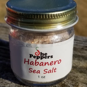 May include: A small glass jar with a gold lid filled with a pink and white sea salt blend. The label reads "The Peppers Habanero Sea Salt 1 oz."