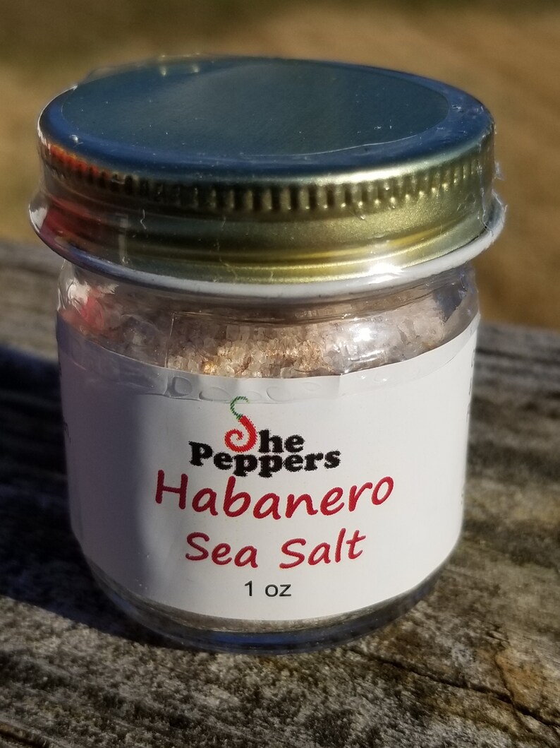 May include: A small glass jar with a gold lid filled with a pink and white sea salt blend. The label reads "The Peppers Habanero Sea Salt 1 oz."
