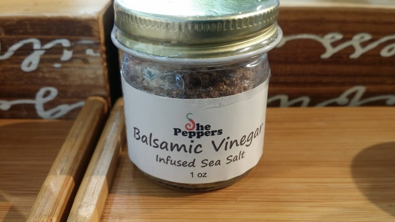 Może przedstawiać: Szklany słoik ze złotą pokrywką wypełniony brązową solą morską infuzowaną octem balsamicznym. Etykieta zawiera napis "The Peppers Balsamic Vinegar Infused Sea Salt 1 oz."
