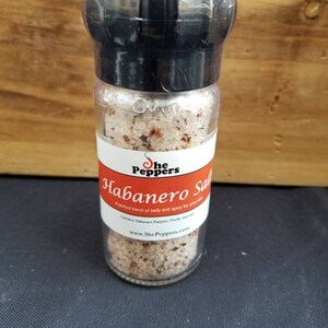 May include: A glass jar with a black lid filled with pink salt and red pepper flakes. The label reads "She Peppers Habanero Salt" and "A perfect blend of salty and spicy for your dish". The website address "www.ShePeppers.com" is printed on the label.