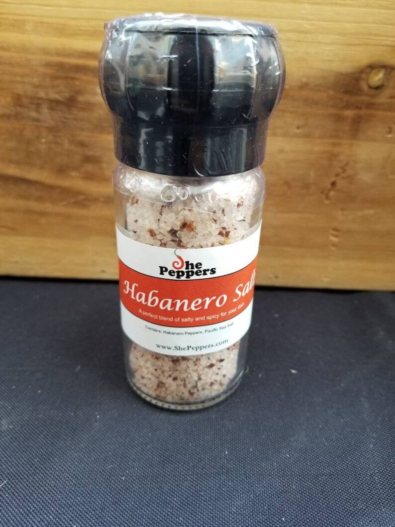 May include: A glass jar with a black lid filled with pink salt and red pepper flakes. The label reads "She Peppers Habanero Salt" and "A perfect blend of salty and spicy for your dish". The website address "www.ShePeppers.com" is printed on the label.