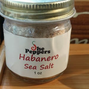May include: A small glass jar with a gold lid filled with a light brown sea salt. The label reads "The Peppers Habanero Sea Salt 1 oz".