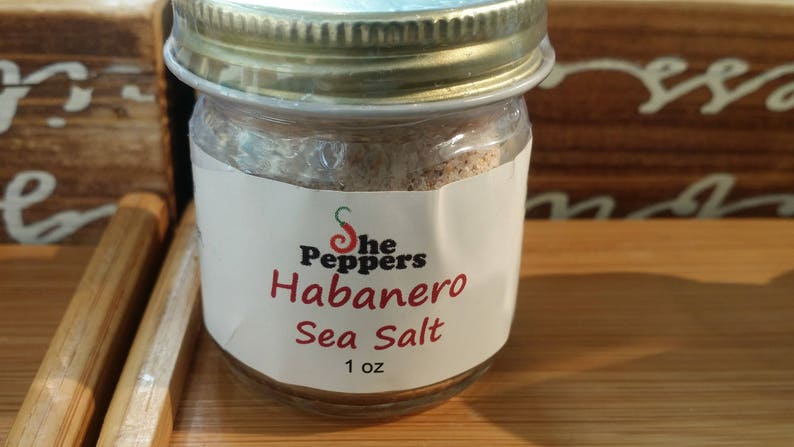 May include: A small glass jar with a gold lid filled with a light brown sea salt. The label reads "The Peppers Habanero Sea Salt 1 oz".