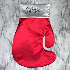 May include: A red satin Christmas stocking shaped like a boxing glove. The top is white satin with the name "Gabriela" embroidered in black. A white satin loop is attached for hanging.