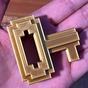 May include: A gold-colored key shaped like a pixelated video game character. The key is made of plastic and has a textured surface.