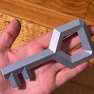 May include: A geometric, silver key with a rectangular handle and a keyhole-shaped opening. The key's teeth are rectangular. The key is held in a hand, with a wooden surface in the background.