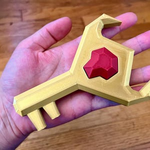 Peut inclure: Une clé dorée stylisée avec une gemme rouge au centre. La clé a un design géométrique unique et est tenue dans une main. Le design de la clé rappelle un objet fantastique ou de jeu vidéo.