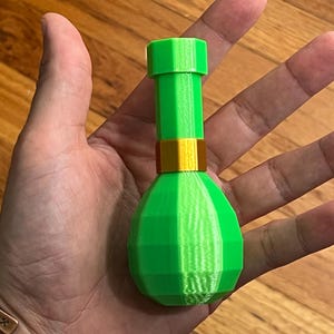 Op de afbeelding: Een groene en gouden fles in de vorm van een toverdrankfles. De fles is gemaakt van plastic en heeft een getextureerd oppervlak.