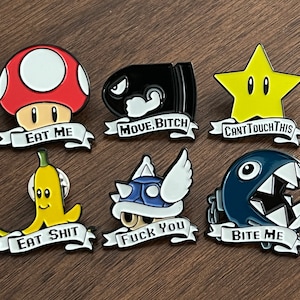 Może przedstawiać: Zestaw 8 emaliowanych pinów z różnymi postaciami z kreskówek i napisami. Piny zawierają czerwonego grzyba z napisem "Eat Me", czarną kulkę z napisem "Move, Bitch", żółtą gwiazdę z napisem "Can't Touch This", żółty banan z napisem "Eat Shit", niebieskiego żółwia z napisem "Fuck You", niebieskiego potwora z napisem "Bite Me" oraz dwa inne piny.