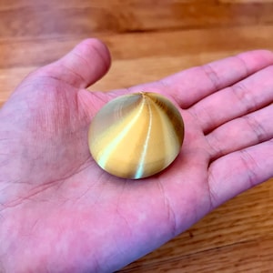 May include: A golden yellow, round, smooth, decorative object with a pointed top and a swirling pattern. The object is being held in a person's hand.