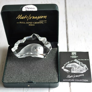 May include: A clear lead crystal sculpture of a polar bear encased in a jagged ice-like form, presented in a dark green hinged box with gold lettering reading Mats Jonasson Full Lead Crystal Sweden, accompanied by a black informational card.