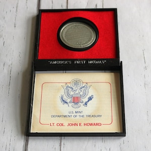 May include: A black box with a red interior holds a silver medal. The box is open, revealing a certificate with the text "AMERICA'S FIRST MEDALS" and the U.S. Mint seal. The certificate also includes the text "DEPARTMENT OF THE TREASURY" and "LT. COL. JOHN E. HOWARD."