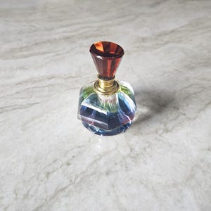 May include: A small, multifaceted glass perfume bottle featuring a clear, iridescent base that reflects blue and green hues. The bottle is topped with a faceted, deep amber-colored glass stopper and a gold-toned metal neck collar.