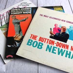 May include: A stack of vintage vinyl records. The album covers feature text and images, including "Here's Johnny," "The Button-Down Mind of Bob Newhart," and "Starring Johnny Carson." The records are on a light wood surface.