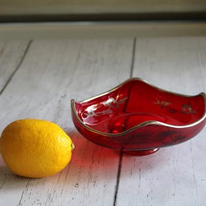 May include: A red glass candy dish with a silver rim and floral design. The dish is on a white wooden surface with a yellow lemon beside it.