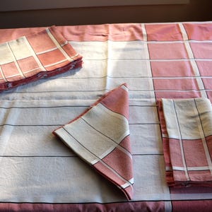Puede incluir: Un mantel de mesa a cuadros rosa y blanco con tres servilletas dobladas. Las servilletas están dobladas de diferentes maneras y el mantel tiene un borde blanco.