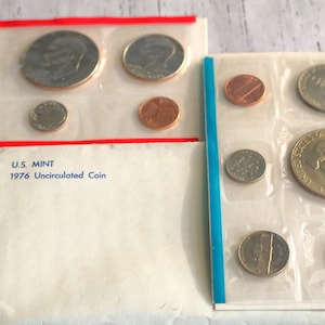 May include: Two coin sets in plastic sleeves, one with a red border and the other with a blue border. The sets contain various U.S. coins, including quarters, dimes, and pennies. A white envelope with the text "U.S. MINT 1976 Uncirculated Coin" is also present.
