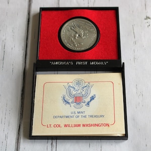 May include: A silver coin in a red and black box with the text "America's First Medals" and "U.S. Mint Department of the Treasury LT. COL. WILLIAM WASHINGTON".