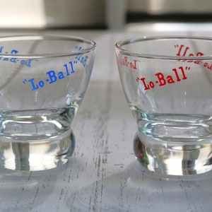 May include: Two clear, lowball glasses with a rounded base. The text "Lo-Ball" is printed on the glasses in blue and red. The glasses are sitting on a white wooden surface.