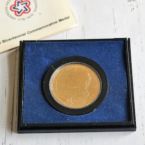 May include: A gold-colored medal in a black display case with a blue velvet lining. The medal features a profile of a person with the text "AMERICAN REVOLUTION 1775-1783" around the edge.