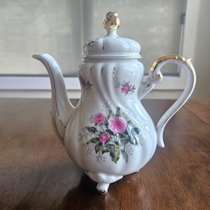 May include: White porcelain teapot with a gold handle, spout, and lid accents. The teapot features a floral design with pink roses and green leaves. The teapot has a decorative lid and sits on three small feet.