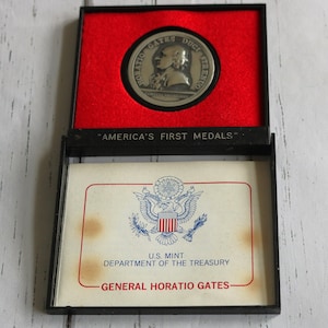 May include: A black display case with a red velvet interior holds a silver medal. The medal features a profile of a person and the words "HORATIO GATES DUCI STRENUO". Below is a card with the text "AMERICA'S FIRST MEDALS".