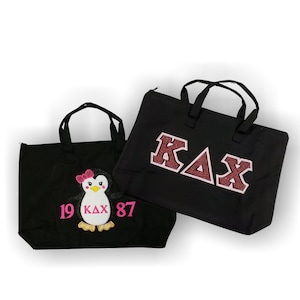 May include: Two black tote bags. One bag features a pink and white penguin with a pink bow and the text "19 KAX 87". The other bag features the letters "KAX" in red glitter.
