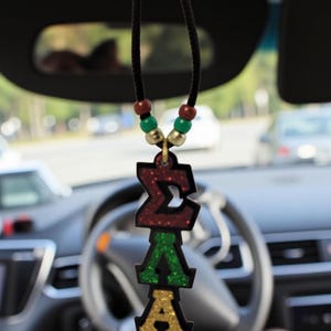 May include: A car air freshener featuring the Greek letters ΣΔΑ in red, green, and gold glitter. It hangs from a black cord with wooden and green beads. The background shows a car interior.