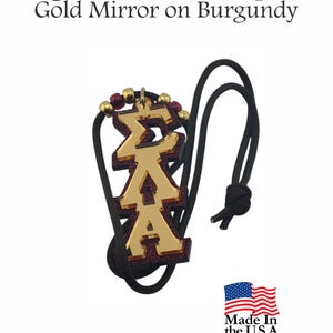 Peut inclure: Un pendentif Sigma Lambda Alpha avec des lettres grecques miroir dorées superposées sur un fond bordeaux à paillettes. Le pendentif est fixé à un cordon noir avec de petites perles rouges et dorées. Le texte indique Sigma Lambda Alpha Gold Mirror on Burgundy.