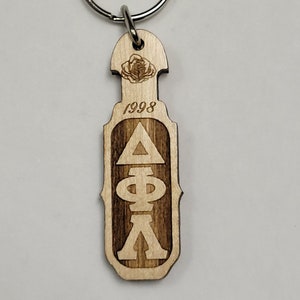 May include: Wooden keychain with a silver ring. The keychain is engraved with the year "1998" and Greek letters. A rose is engraved at the top. The keychain is made of light-coloured wood.