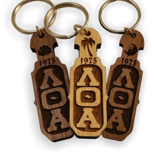May include: Three wooden keyrings with a gold key ring. Each keyring features a different design with the letters "AOA" and the year "1975" engraved. The designs include a seashell, a palm tree, and a crescent moon.