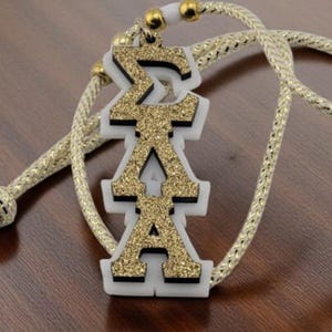 May include: A gold glitter pendant with the Greek letters ΣΑΑ, on a gold rope necklace. The pendant has a white border and is set against a dark wood surface. The necklace has gold accents.