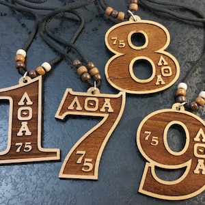 May include: Four wooden necklaces with pendants shaped like the numbers 1, 7, 8, and 9. Each pendant has the letters "AOA" and the number "75" engraved on it.