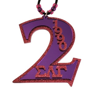 May include: Purple and red glitter number 2 pendant with the text "1990" and "ΣΛΓ" on a black cord with pink and purple beads.