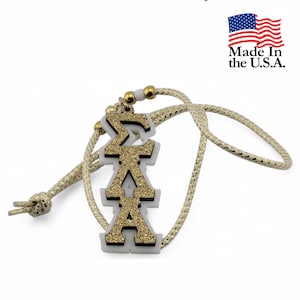 May include: A gold glitter pendant featuring the Greek letters Sigma, Lambda, and Alpha on a white acrylic base, suspended from a gold cord. The text "Sigma Lambda Alpha" is at the top.