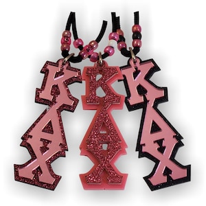 May include: Three pink and black sorority keychains with the letters "KAX" in a bold, stylised font. The middle keychain has glitter on the letters.