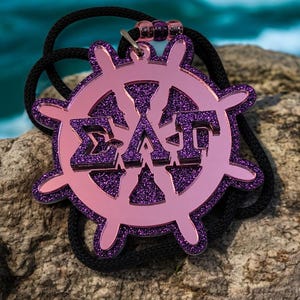 May include: A pink and purple pendant necklace featuring a ship's wheel design with Greek letters. The pendant has a glittery purple center and a pink outline. The necklace has a black cord.