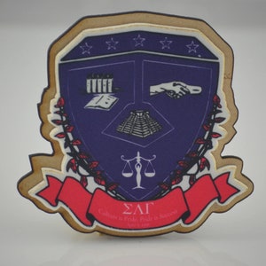 Peut inclure: Un bouclier en bois avec un centre violet et une bordure dorée. Le bouclier présente un livre, une pyramide, une main, une balance et un ruban rouge avec le texte "ΣΛΓ Culture is Pride, Pride is Success 1911-1991".