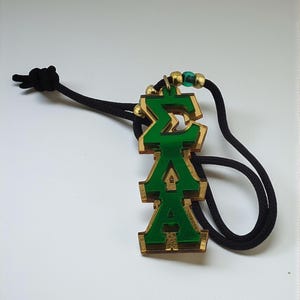 May include: A green and gold pendant necklace featuring the Greek letters Sigma, Xi, and Alpha. The pendant hangs from a black cord. The necklace has gold beads and a green bead near the clasp.