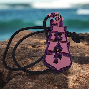 May include: A pink and purple pendant with a black cord. The pendant has a layered design with a glittery purple border. The background shows a rocky surface and the ocean.