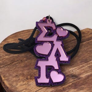 May include: A purple and pink pendant necklace with the Greek letters ΣΛΓ, accented with glitter and heart shapes. The pendant hangs on a black cord, resting on a wooden surface.