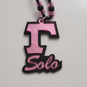 May include: A pink and black necklace with a pendant that says "T Solo". The pendant is made of acrylic and has a mirrored finish.