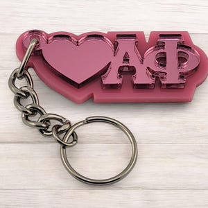 May include: Pink acrylic keychain with a heart and the Greek letters ΑΦ. The keychain has a silver-colored chain and a keyring. The letters and heart are a reflective, mirrored finish.