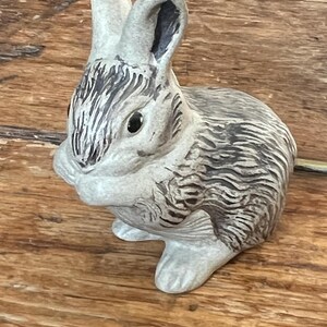 May include: A ceramic rabbit figurine in a seated pose, with a light grey and brown colour scheme. The rabbit has detailed fur texture and black eyes. The figurine is on a wooden surface.