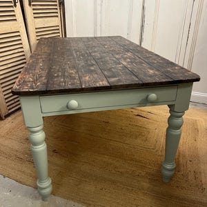 May include: A rectangular wooden table with a dark brown, plank-style top and a light green base. The base features a drawer with two round knobs and four turned legs. The table is approximately 122 cm long.