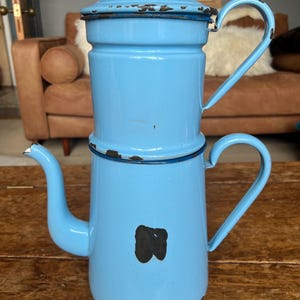 May include: A vintage, light blue enamel coffee pot with a spout, handle, and lid. The pot has a distressed appearance with dark brown spots and a black mark on the front. The pot is sitting on a wooden surface.