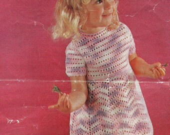 May include: A young girl wearing a pink and white crocheted dress, holding a small green leaf in her hand. She is standing in front of a pink background and a wooden wagon filled with purple and white flowers.