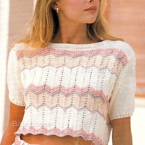 May include: A white and pink knit crop top with short sleeves. The top features a scalloped design in white, pink, and light pink. The model is wearing a white skirt and a pink belt.