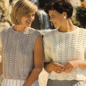 Puede incluir: Dos mujeres con tops de punto. Una lleva un top sin mangas gris claro con un patrón de vieira, combinado con una falda blanca. La otra lleva un top crema de manga corta con un diseño similar. Los tops son de Babbabees.